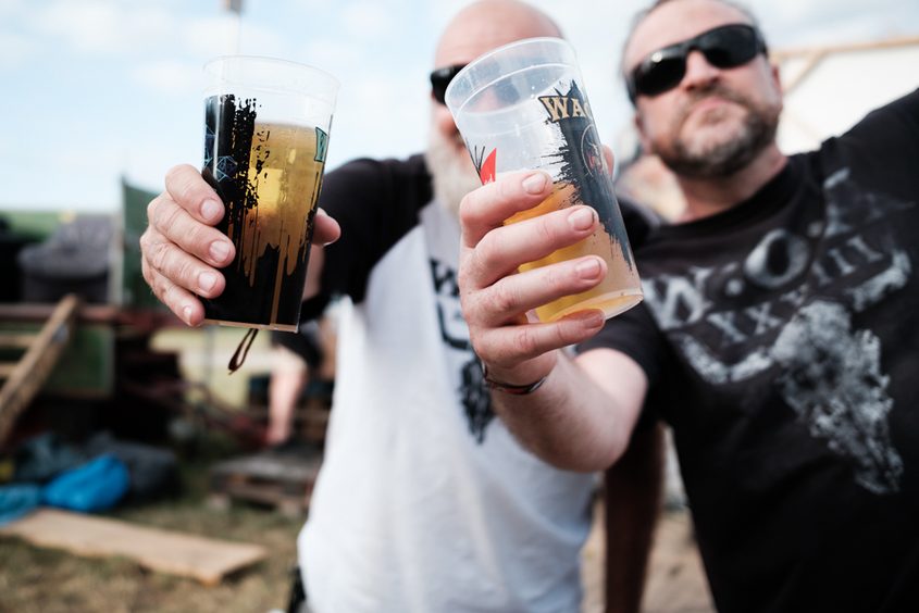 Konzert-Reportage beim Wacken Open Air: Zwei Metalfans prosten sich lachend mit Bierdosen auf dem staubigen Campground zu.
