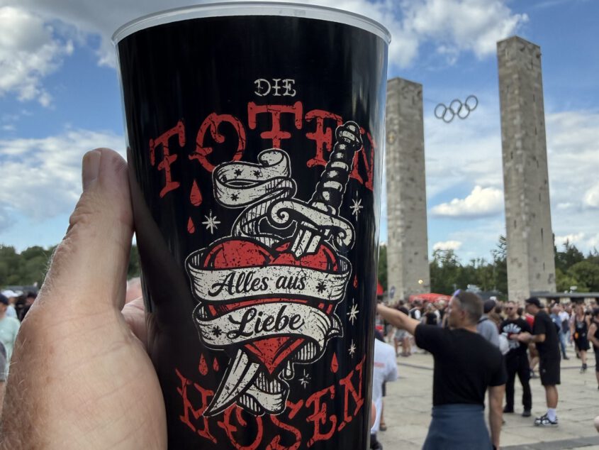 Eventreportage vor einem Stadion: Ein hochgehaltener, bedruckter Becher der Band Die Toten Hosen, fotografiert bei einer überregionalen Konzert-Dokumentation.