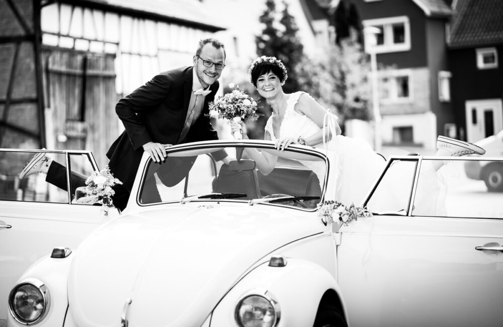 Brautpaar im weißen Käfer-Cabrio mit Blumenschmuck auf dem Weg zur Feier in Hößlinsülz