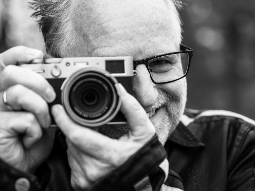 Fotograf Peter Goedecke: Schwarz-weißes Portrait des Fotografen mit Kamera vor dem Gesicht an einer Outdoor-Location, überregional und europaweit im Einsatz.
