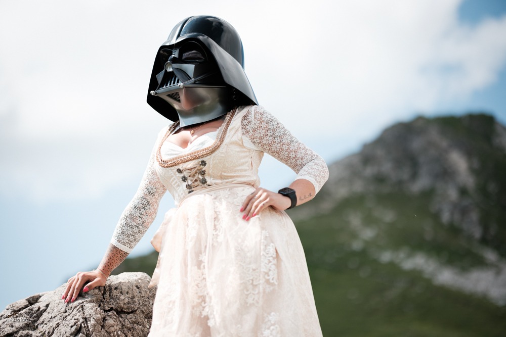 Frau im hellen Dirndl mit Darth-Vader-Maske auf einem Felsen, dahinter der Grat der Allgäuer Berge