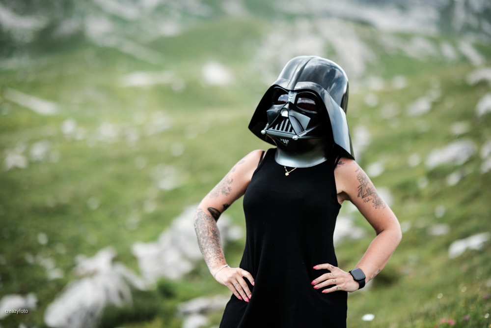 Frau im schwarzen Sommerkleid mit Darth-Vader-Maske, die Hände in die Hüften gestemmt, dahinter unscharf eine Almwiese in den Bergen über Oberstdorf.