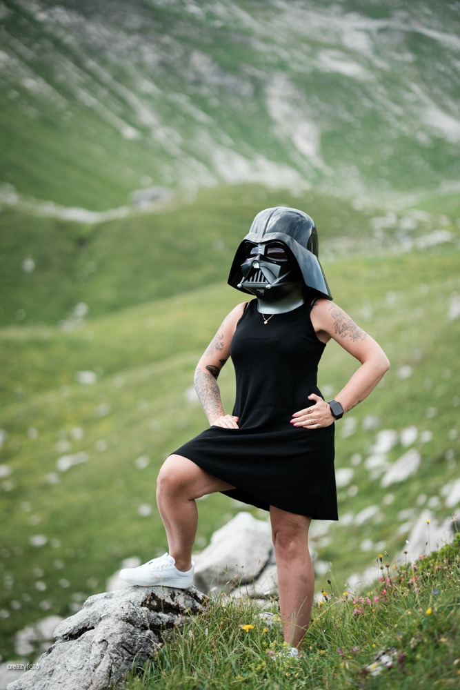 Ganzkörperaufnahme einer Frau mit Darth-Vader-Maske im schwarzen Kleid, ein Fuß auf einem Felsblock, im Rücken die grüne Bergflanke über Oberstdorf.