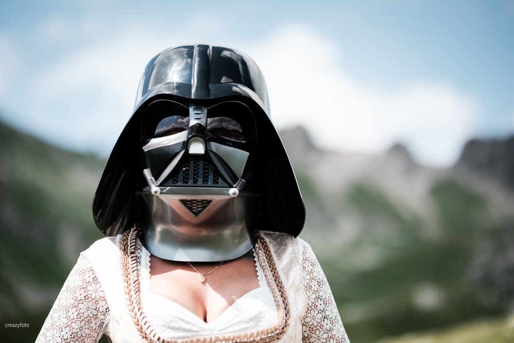 Nahaufnahme der Darth-Vader-Maske über dem Spitzenmieder eines Dirndls, im Hintergrund die Berge