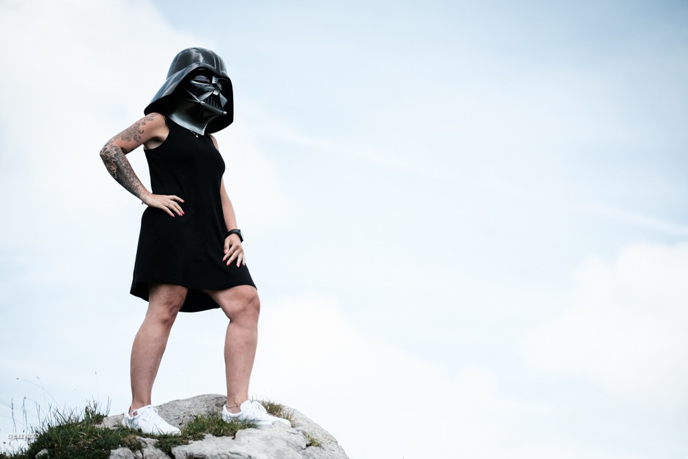 Frau mit Darth-Vader-Maske steht auf einer Felskuppe gegen den offenen Himmel, neben ihr nur Wolken.