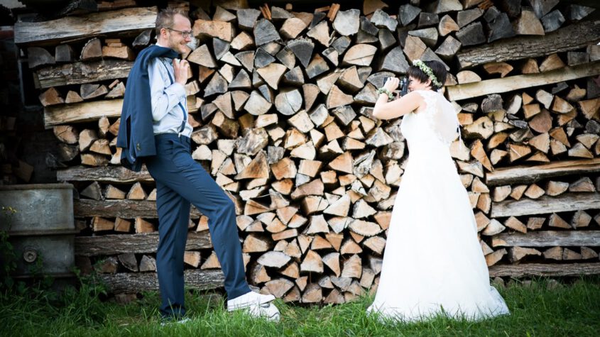 Braut fotografiert ihren lachenden Bräutigam vor einem Holzstapel, er posiert übertrieben