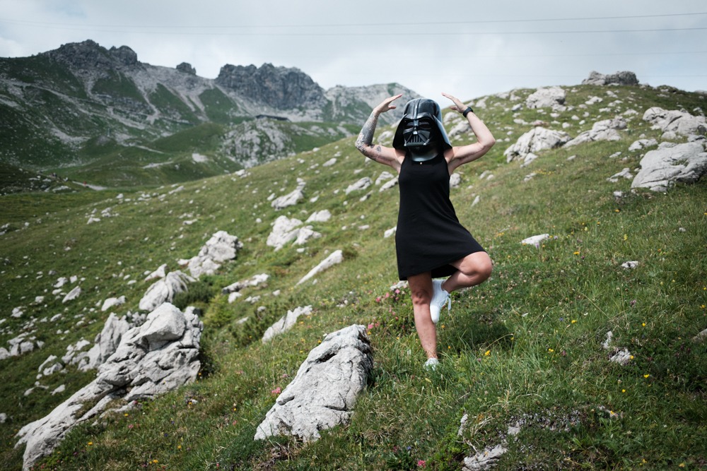 Frau mit Darth-Vader-Maske reckt die Arme über den Kopf, vor ihr eine Blumenwiese mit Felsblöcken, dahinter die Gratkette der Allgäuer Alpen.