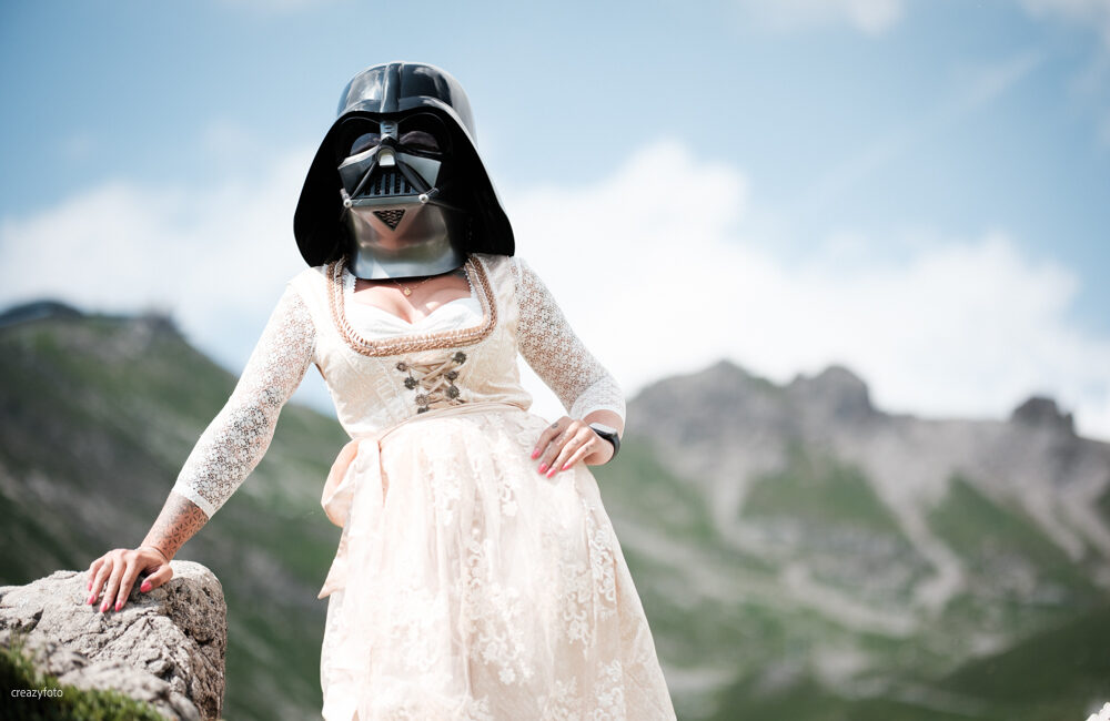 Frau im hellen Spitzendirndl mit Darth-Vader-Maske im hellen Mittagslicht, hinter ihr die Bergkette über Oberstdorf.