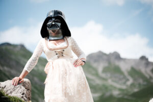 Frau im hellen Spitzendirndl mit Darth-Vader-Maske im hellen Mittagslicht, hinter ihr die Bergkette über Oberstdorf.