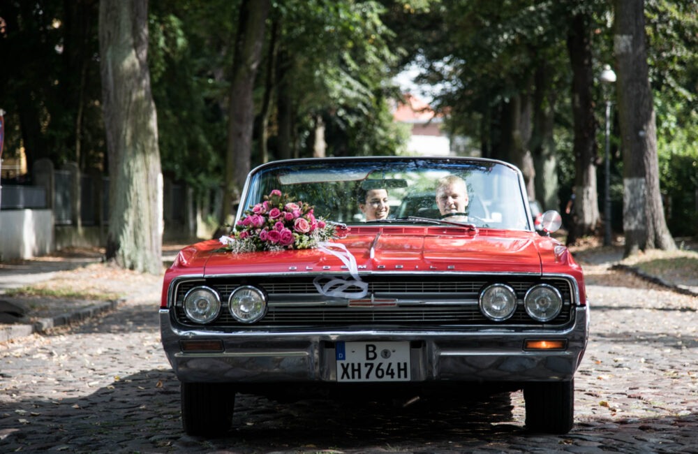 Das rote Oldsmobile-Cabrio steht frontal in einer Baumallee bei Schloss Glienicke, Sarah und Timo sitzen darin.