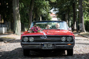 Das rote Oldsmobile-Cabrio steht frontal in einer Baumallee bei Schloss Glienicke, Sarah und Timo sitzen darin.