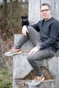 Mann sitzt auf Baumstumpf im Wald mit Hund im Hintergrund, lockeres Portrait – Fotograf aus Heilbronn