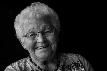 Schwarz-weiß-Portrait einer fröhlichen Seniorin mit Brille und weißen Locken im Fensterlicht – Projekt Lebensspuren, Fotograf Heilbronn-Allgäu-Hamburg-Bremen