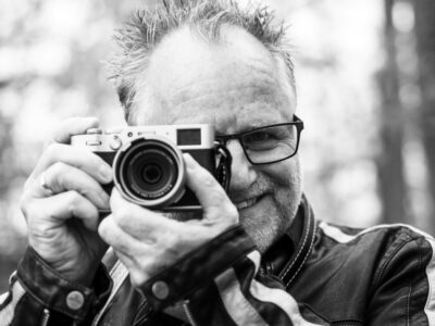 Peter Gödecke mit der Kamera am Auge im Grünen, Porträt von Andrea Kopp