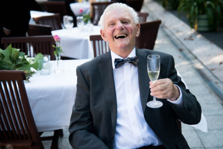 Ein Hochzeitsgast lacht mit dem Sektglas in der Hand an der gedeckten Tafel.